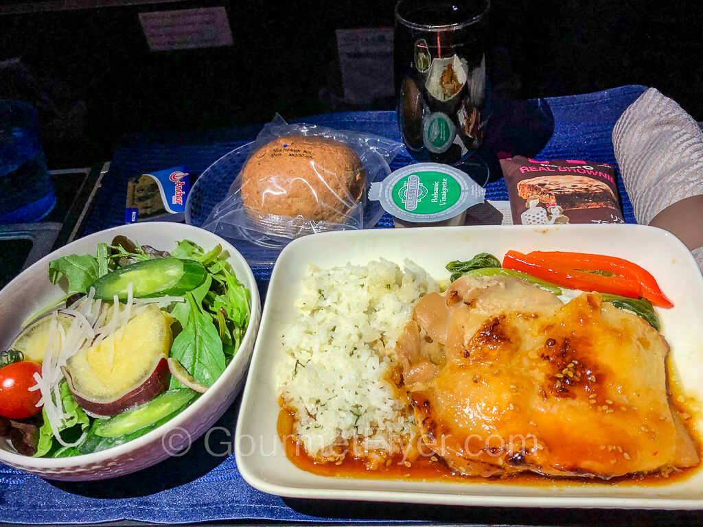 A dinner tray contains a plate of chicken with rice, a green salad, a roll wrapped in plastic, and a glass of red wine.