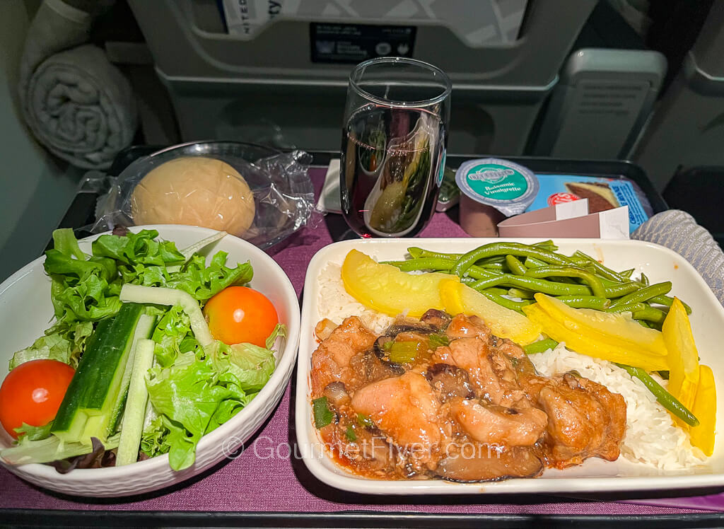 An airliner meal consists of a chicken dish accompanied by salad, bread, a glass of red wine, and dessert.