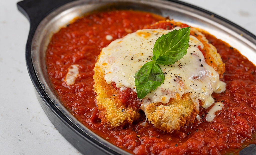 A fried chicken breast filet topped with cheese is served in a metal dish filled with tomato sauce.