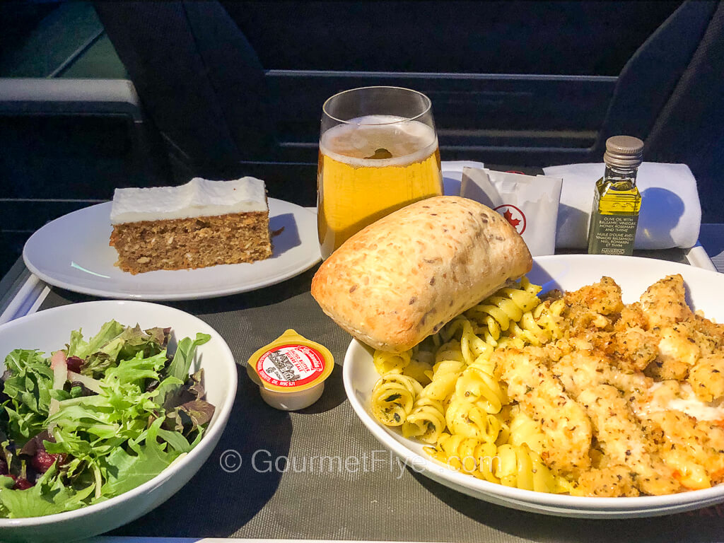 An airplane dinner tray contains a pasta dish accompanied by a green salad, a slice of cake, and a glass of beer.