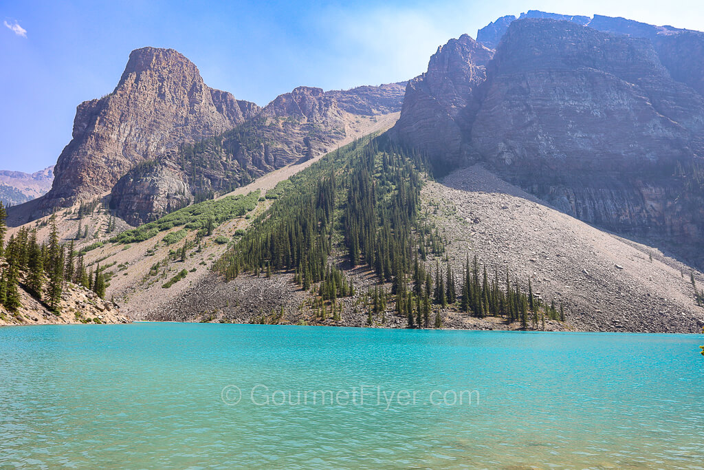 A lake with pristine turquoise color water has rocky mountains with alpine trees in its background.