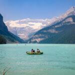 A canoe with two people sails on a lake with turquoise waters under the backdrop of a glacier mountain.