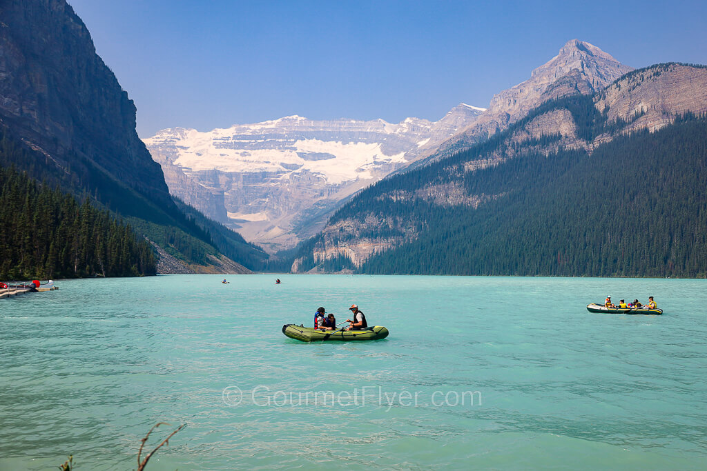 A canoe with two people sails on a lake with turquoise waters under the backdrop of a glacier mountain.