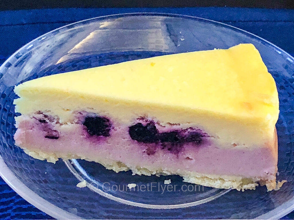 A slice of cheesecake with a thick yellowish cheesy topping and a purple filling punctuated with pieces of blueberries.