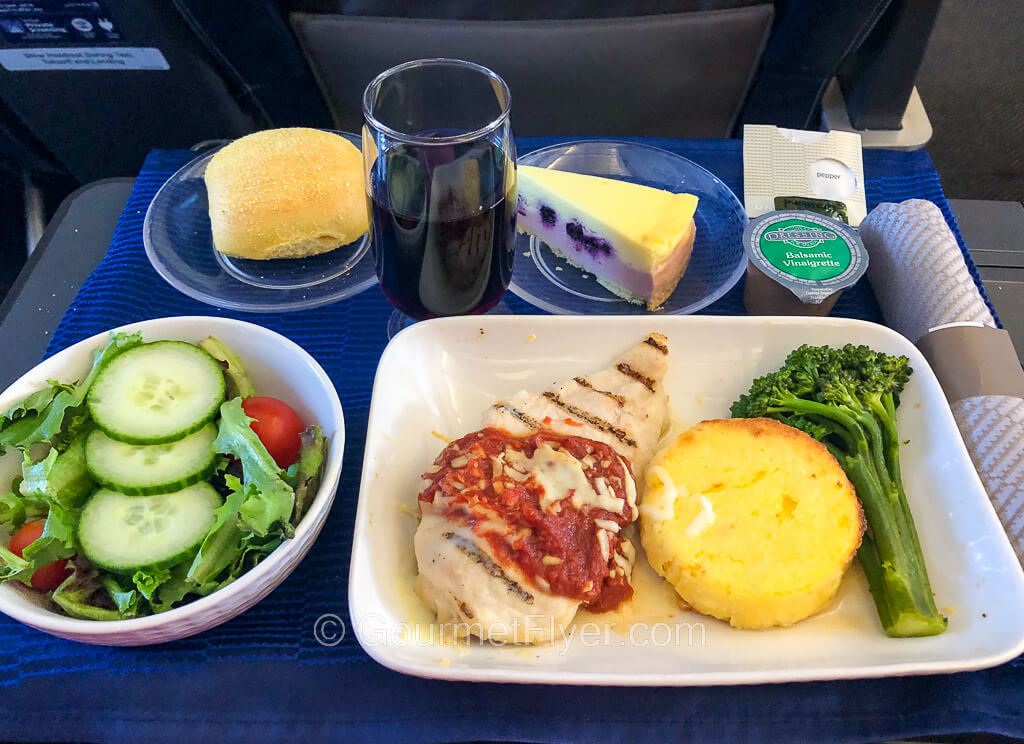 A dinner tray contains a dish with a chicken breast topped with a red sauce, a disc of potato-like pastry, and a broccolini. Side dishes include a salad, bread, and dessert.