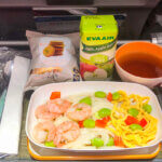 A meal tray carries a dish of shrimp and noodles accompanied by a carton of apple juice and a cup of black tea.