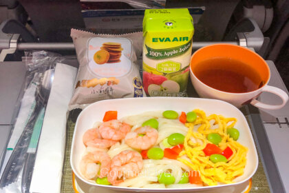 A meal tray carries a dish of shrimp and noodles accompanied by a carton of apple juice and a cup of black tea.