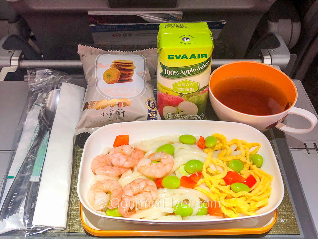 A meal tray carries a dish of shrimp and noodles accompanied by a carton of apple juice and a cup of black tea.