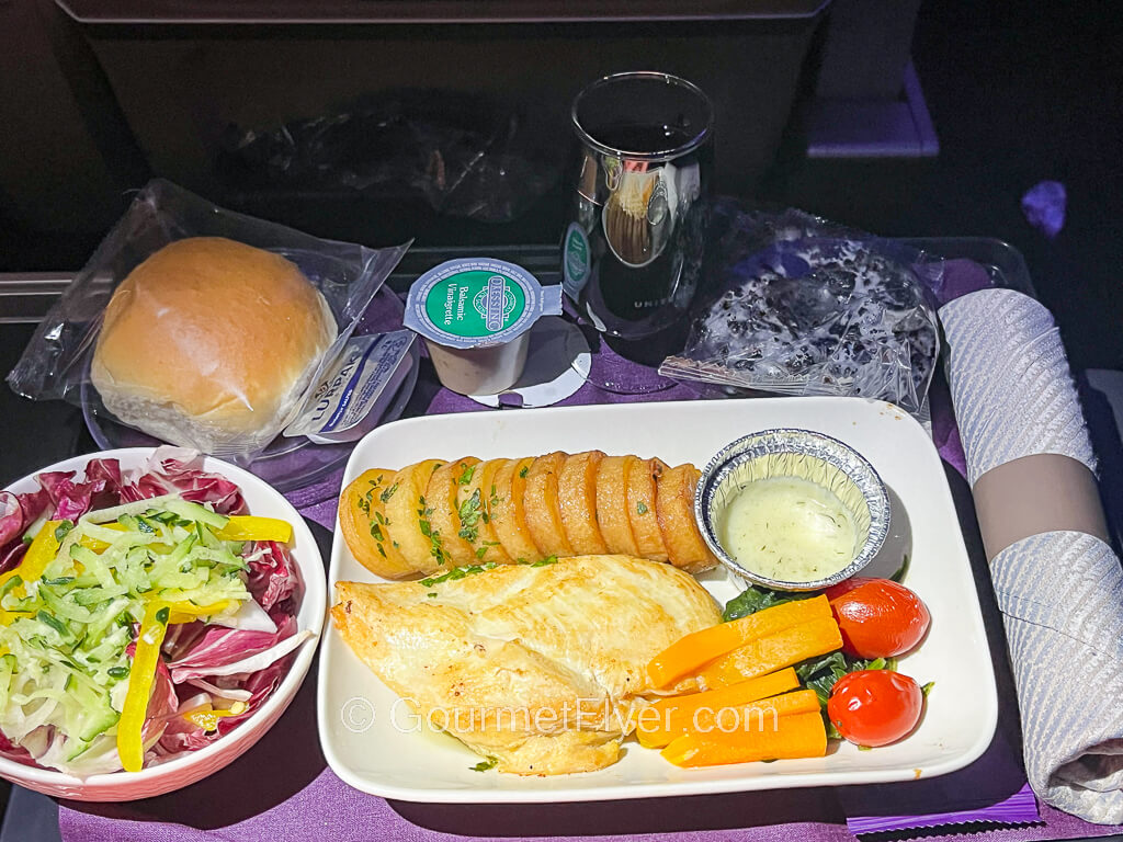 A dinner tray contains a dish of chicken with vegetables, salad, a glass of red wine, and silverware rollup.