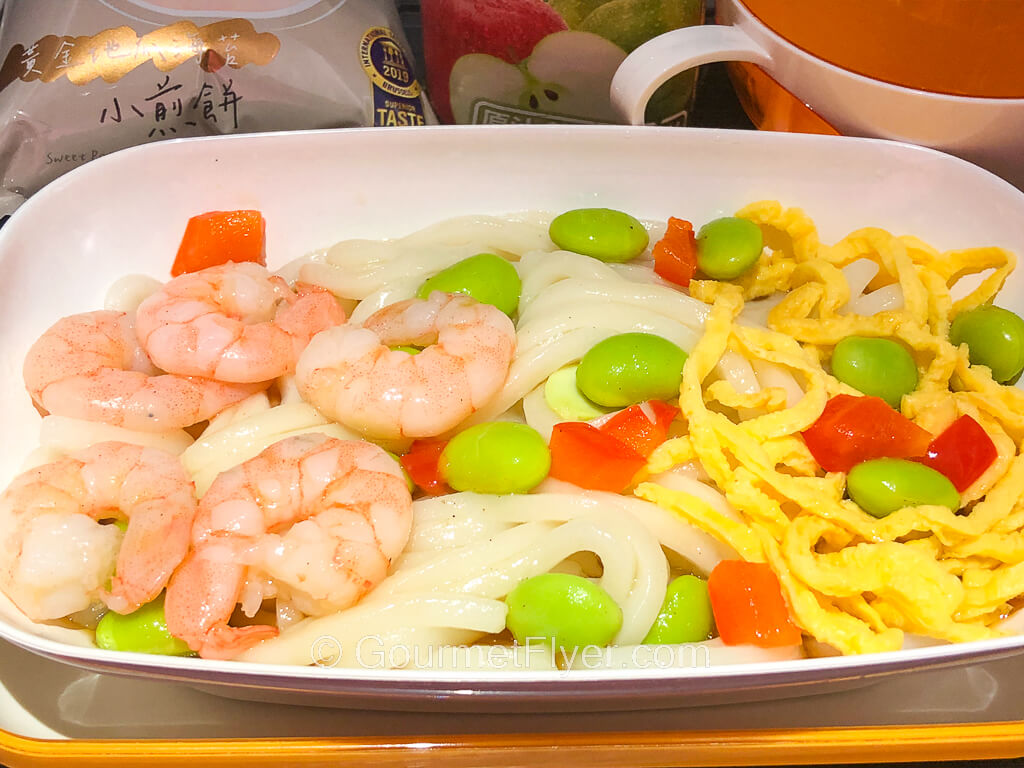 A plate of shrimps and white rice noodles garnished with beans and diced red peppers.