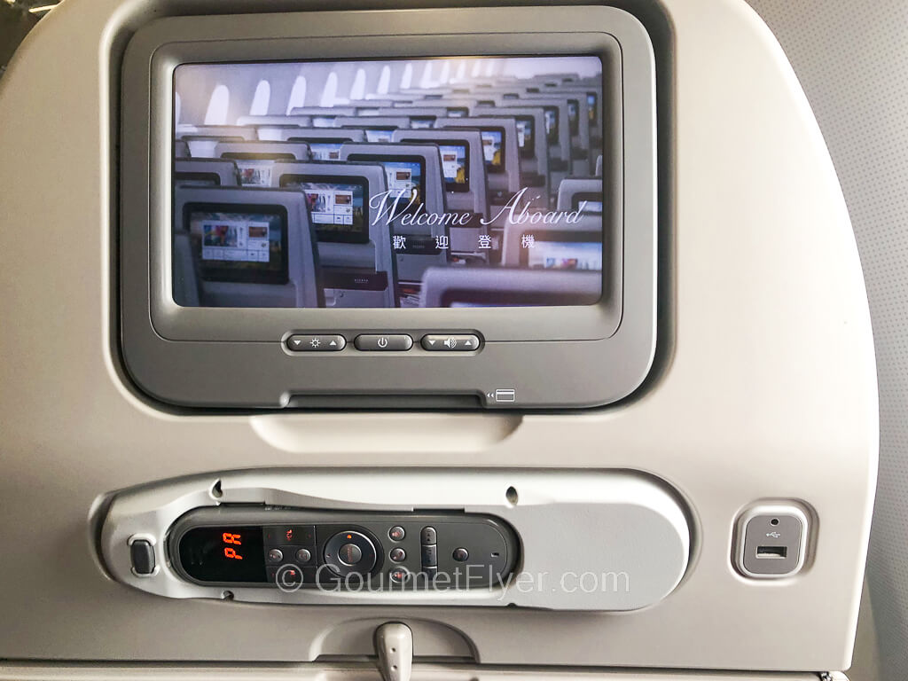 A TV screen is mounted on an airliner's seat back with a remote control in a bracket below it.