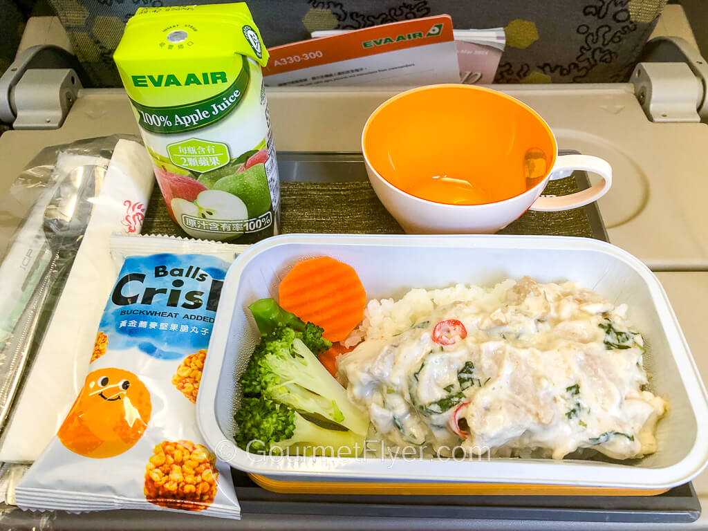 A creamy entree in a dish served with vegetables is accompanied by a box of juice and a pack of crackers.