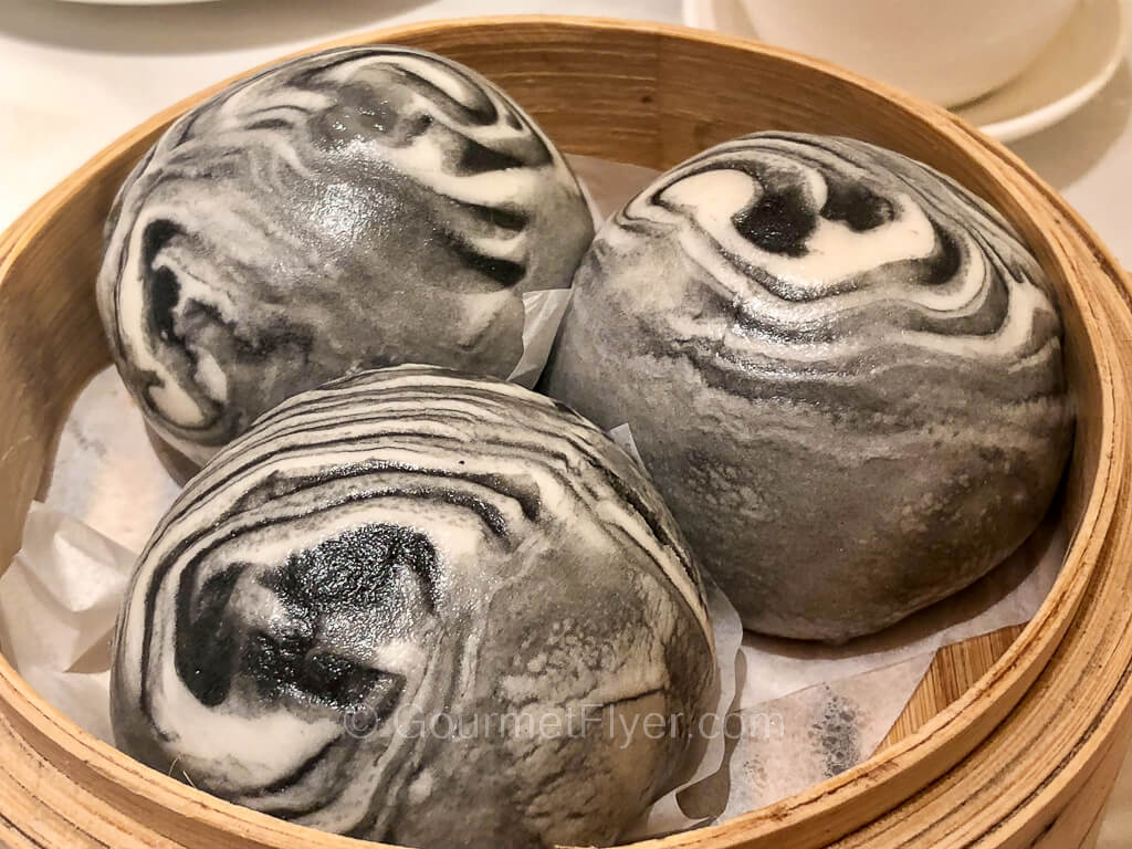 A bamboo dim sum steamer contains three steamed buns with a black and white zebra-looking dough.