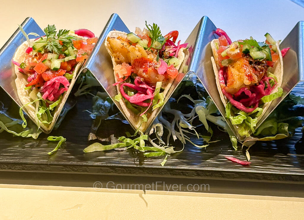 Three tacos with seafood and garnished with colorful vegetables are served on a rack.