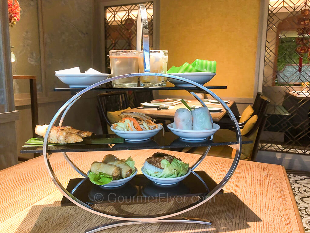 A British style three-tiered afternoon tea platter contains a variety of foods in white small plates and mini glasses.