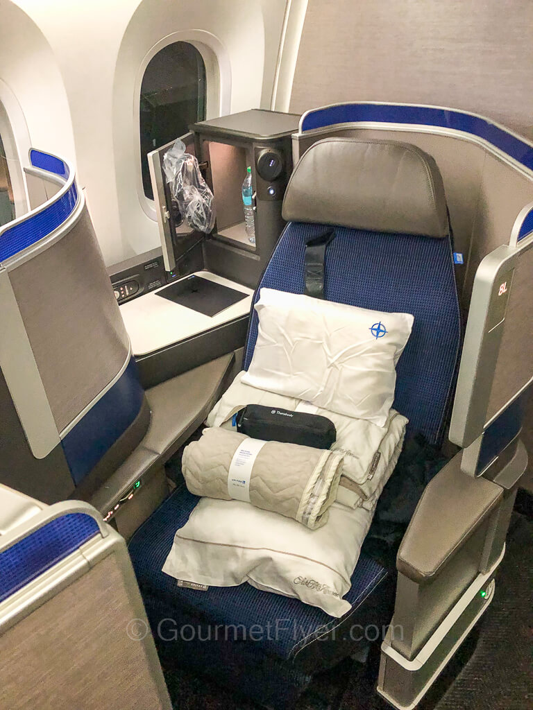 A lie-flat business class seat by the window in its upright position with amenities and beddings placed on top of it.