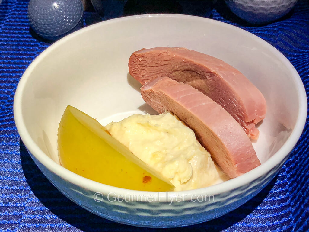 A white porcelain bowl contains two pieces of meat alongside a wedge of apple, served atop a blue tablecloth.