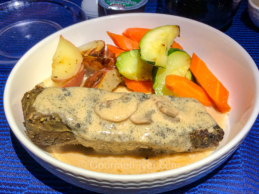 A steak covered in a light color gravy is served with a blend of vegetables in a while porcelain bowl.