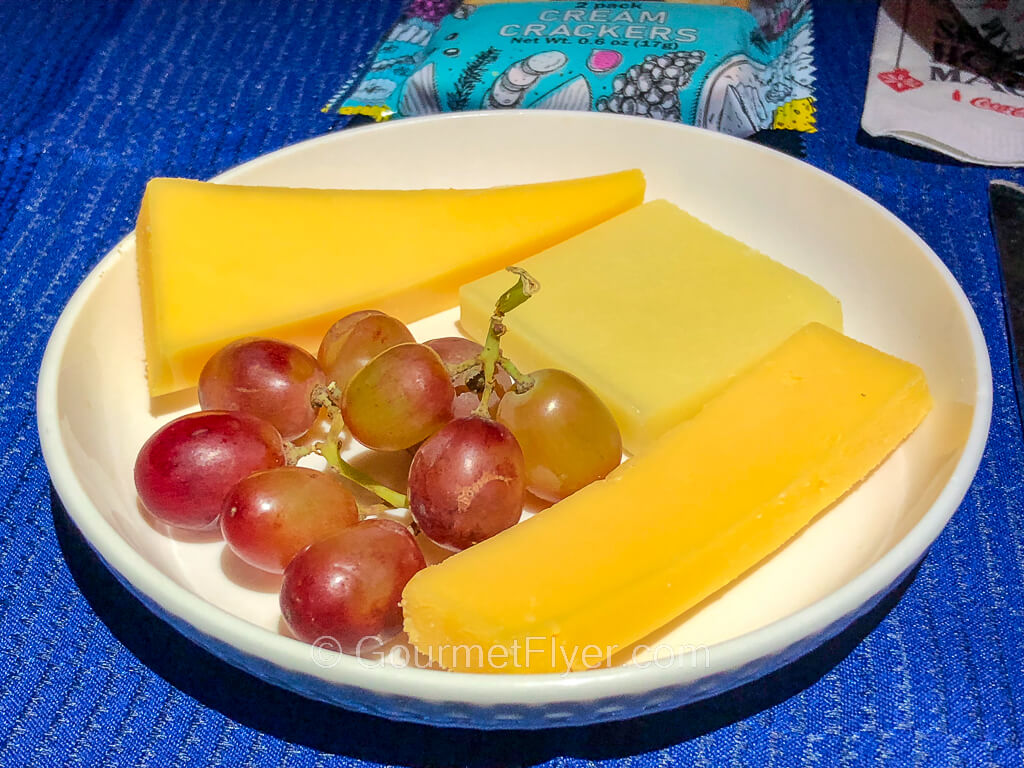 A small white plate contains three pieces of different cheeses and several grapes.