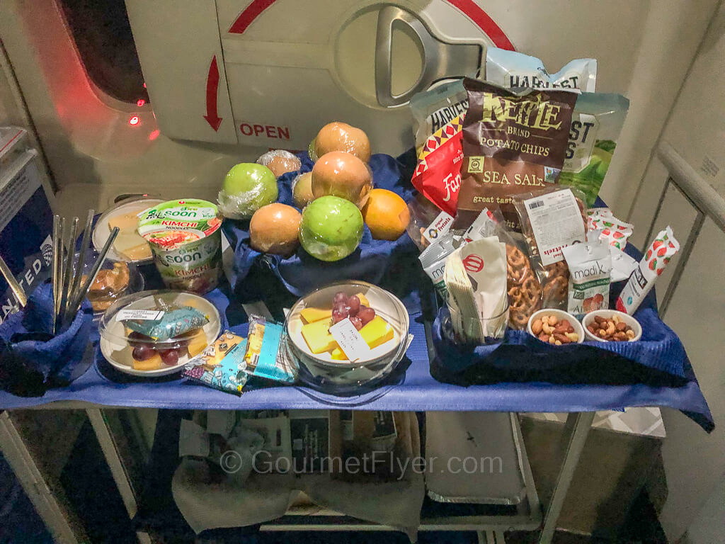 A snack cart has plenty of food piled on top of it, including fruits, packaged snacks, and cheese plates.