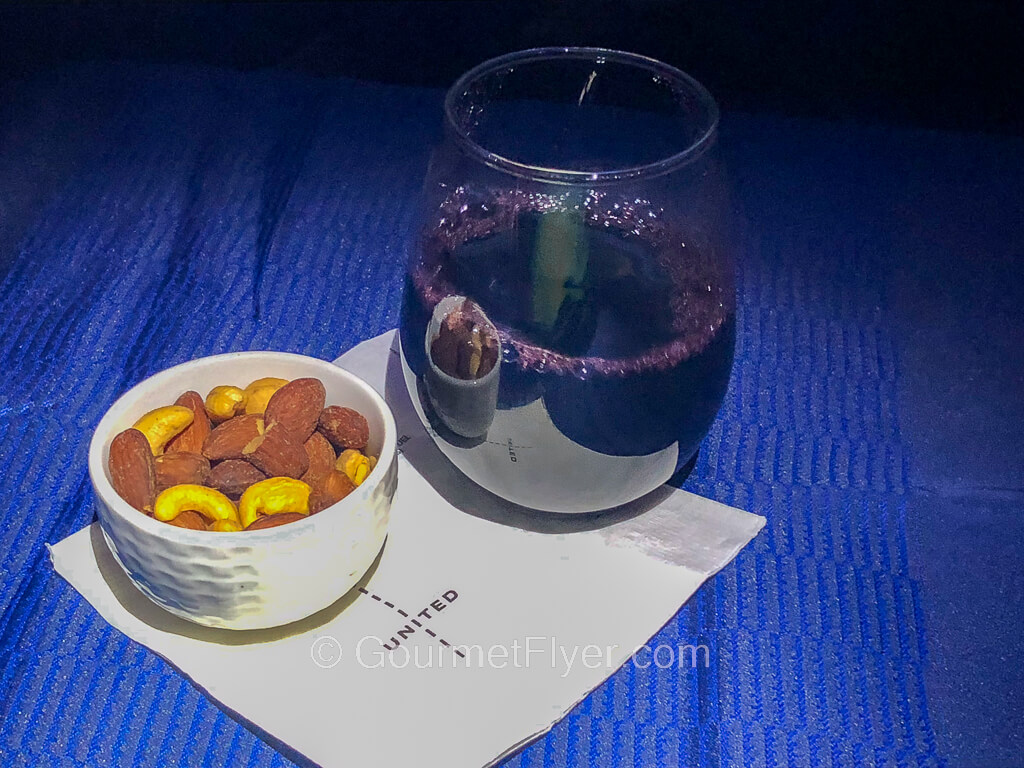 A glass of red wine alongside a ramekin of nuts are served on a napkin atop a blue tablecloth.