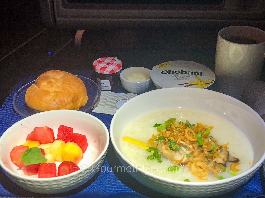 An airline dinner tray contains a bowl of congee accompanied with a bowl of fruits and a pastry.