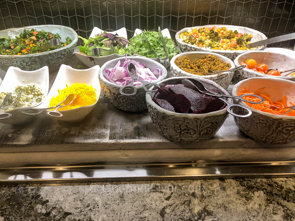 Many small bowls on a buffet counter contains various vegetables and other ingredients of a salad bar.