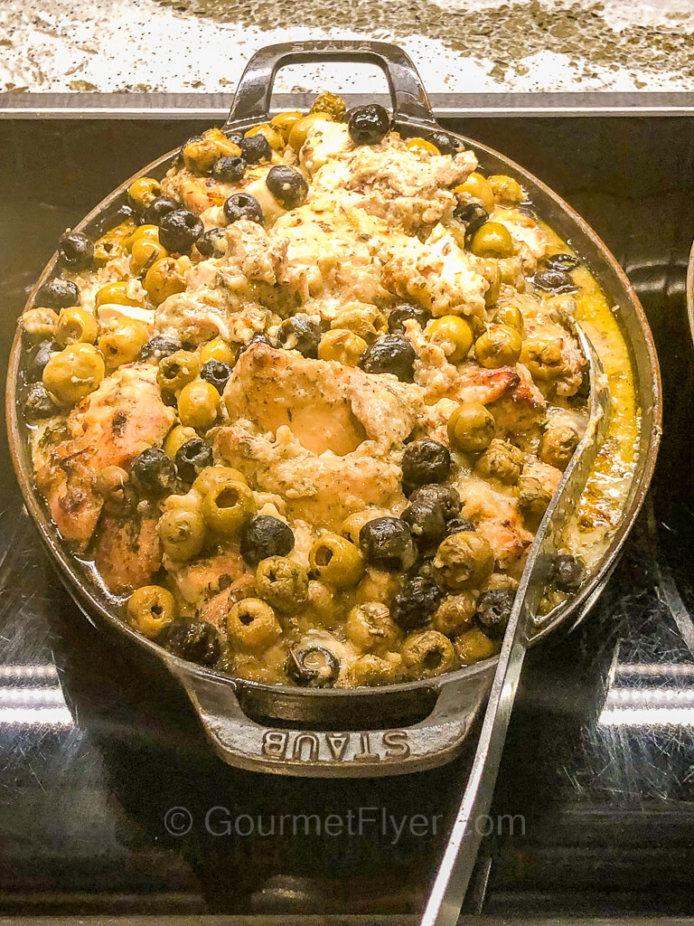 A large metal serving platter is filled to the brim with chicken in a creamy sauce blended with plenty of olives.