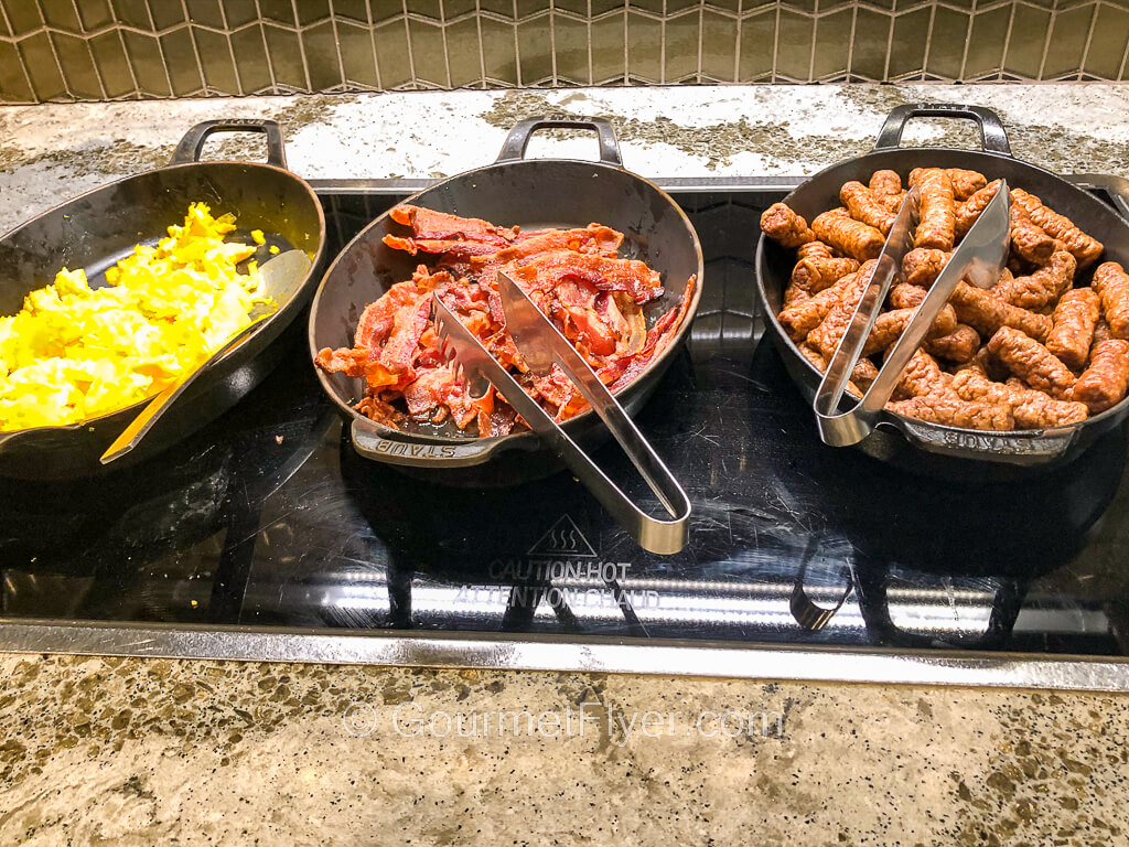 Three serving platters at a buffet contain eggs, bacon, and sausages from left to right.