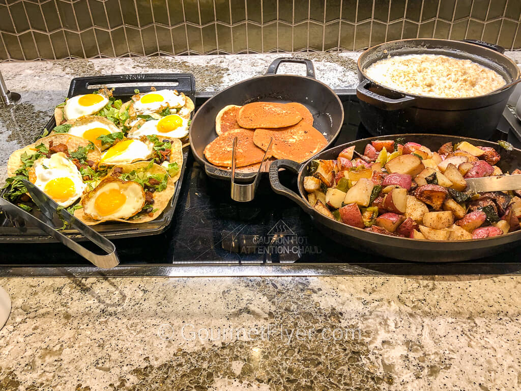 Platters on a buffet counter include a dish with sunny side up eggs, pancakes, and breakfast potatoes.