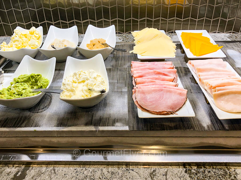 Many small bowls and plates are laid out on a buffet counter containing oatmeal, cold cuts, and cheeses.