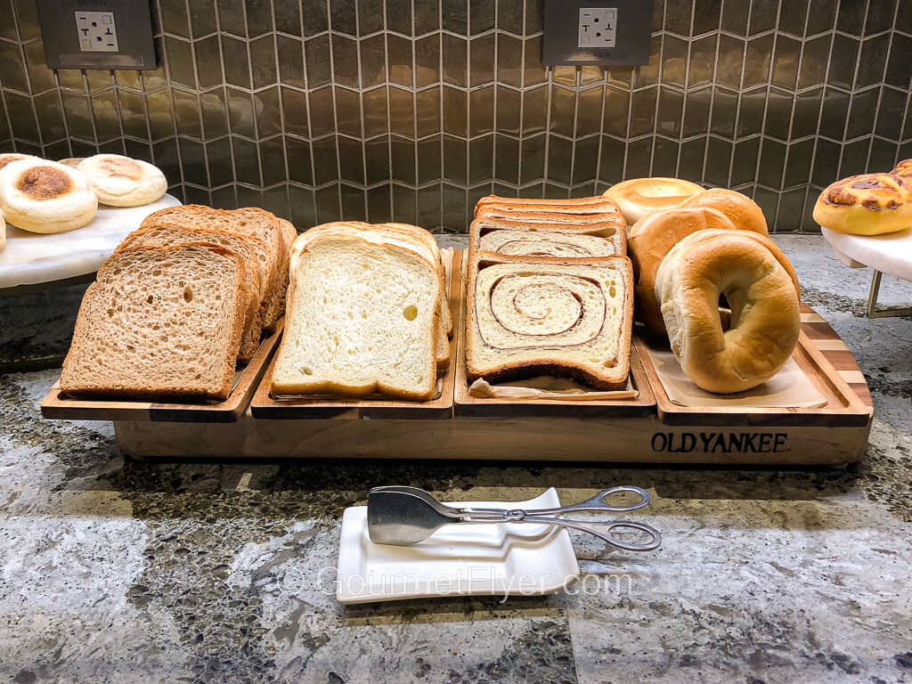 Piles of breads and pastries on wooden serving boards are laid out on top of a buffet counter.