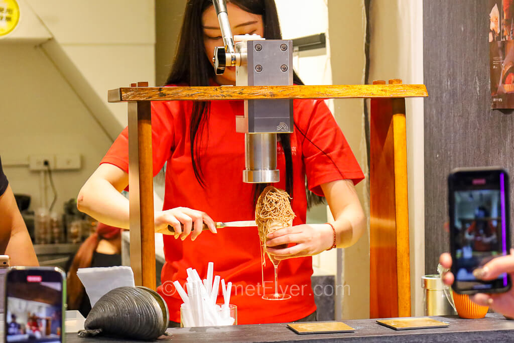 A female server is hand crafting a dessert in a glass under a machine.