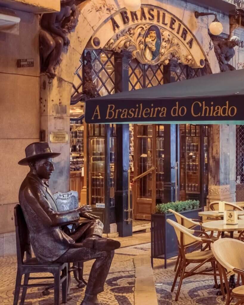 A bronze statue of a man wearing a hat and sitting at a table is placed next to a grand entrance.