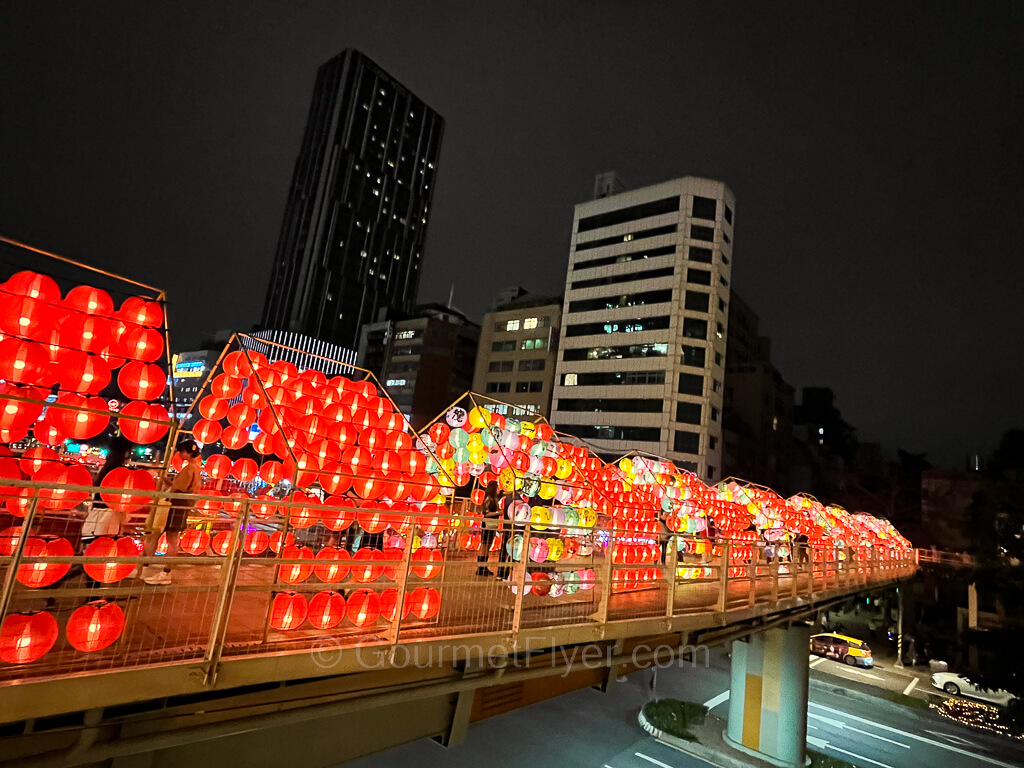 A pedestrian bridge is covered by numerous colorful lanterns which are lit brightly at night.