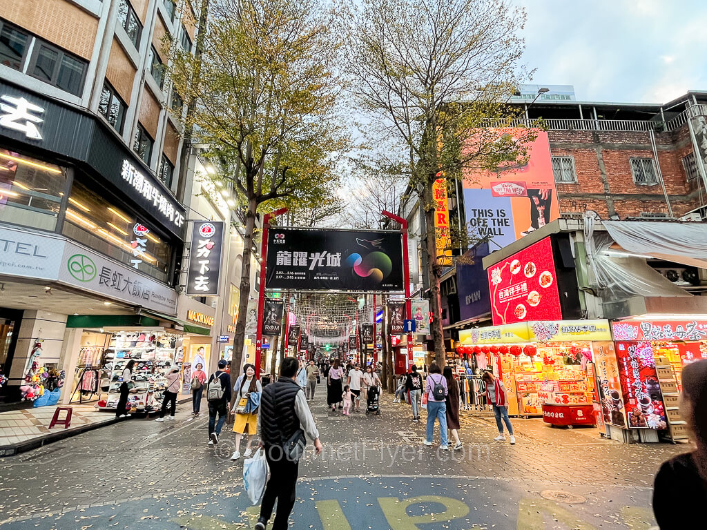 Shoppers stroll down a shopping street lined with brightly lit and colorful stores on both sides.