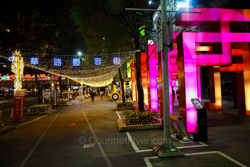 A wide pedestrian sidewalk is decorated with colorful lighted displays.