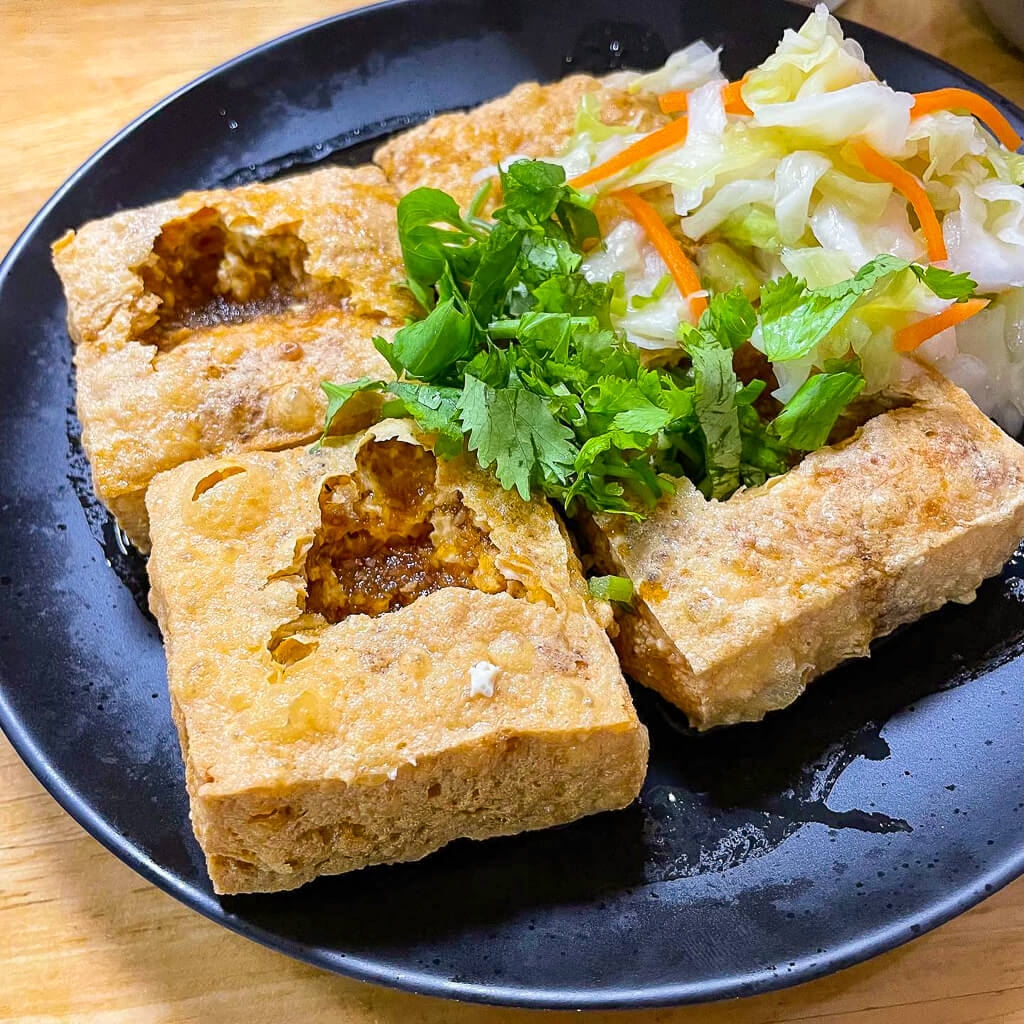 Four square pieces of fried tofu on a plate are topped with parsley and vegetables.