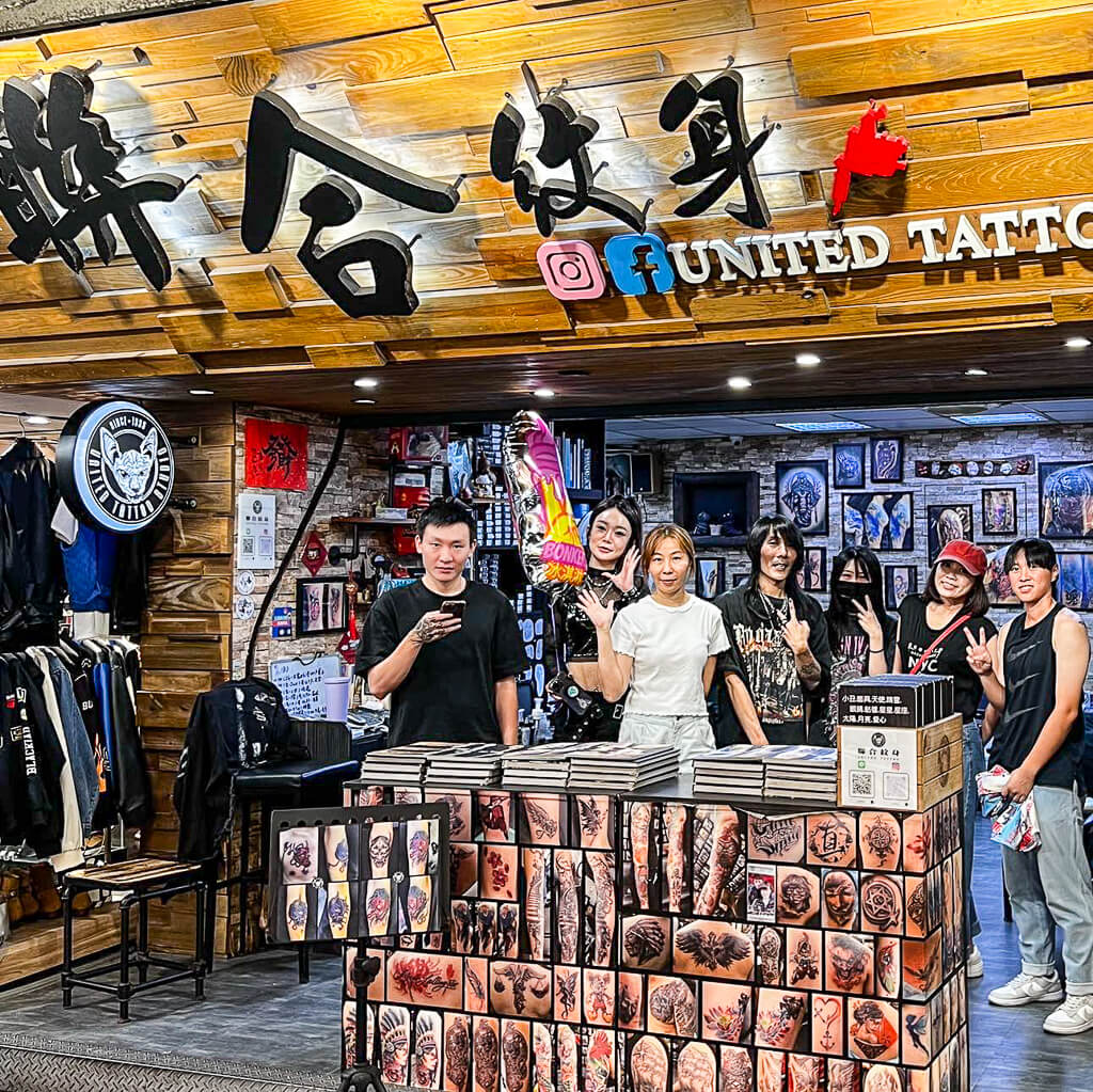 A team of tattoo artists are standing behind a wall of pictured samples and waving to the camera.