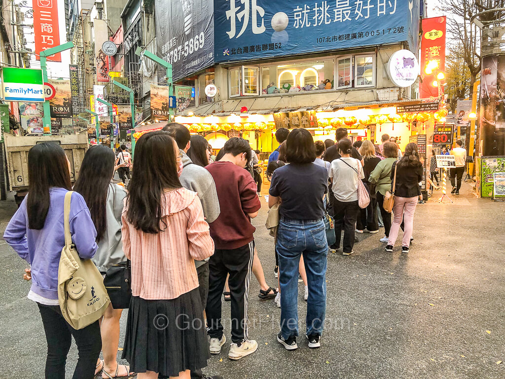 A long line of 20 to 30 people forms on a street, waiting to enter a store.