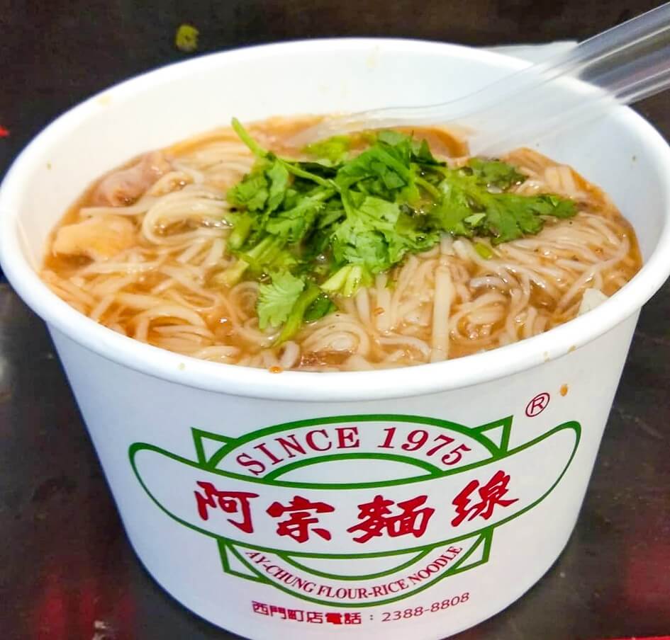 A paper bowl printed with a restaurant's logo contains noodle soup topped with a sprinkle of parsley.