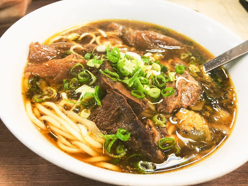 A bowl of noodle soup is topped with sliced beef and sprinkled with green onions.