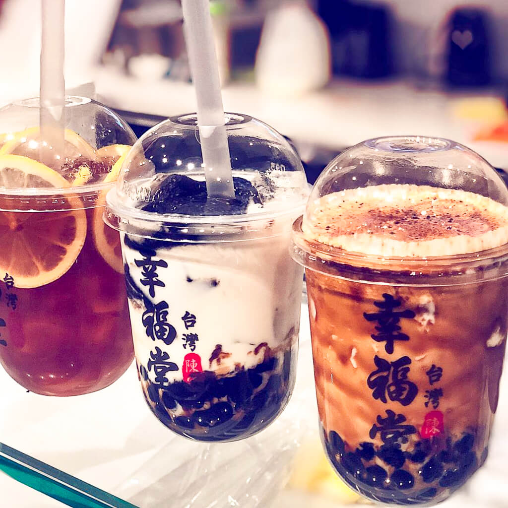 Three different boba drinks in plastic cups from left to right are lemon iced tea, a milky drink, and a coffee like drink. 