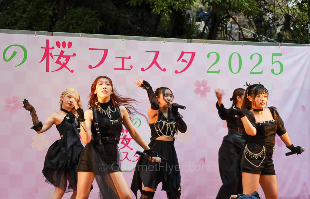 A group of five girls wearing black skirts are singing and dancing on stage.