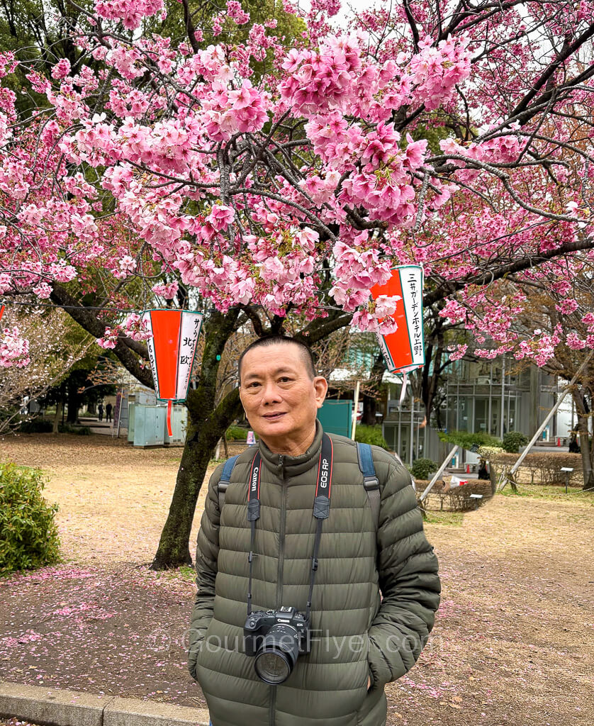 The blogger Gourmet Flyer poses in front of a cherry blossom tree with pink flowers.