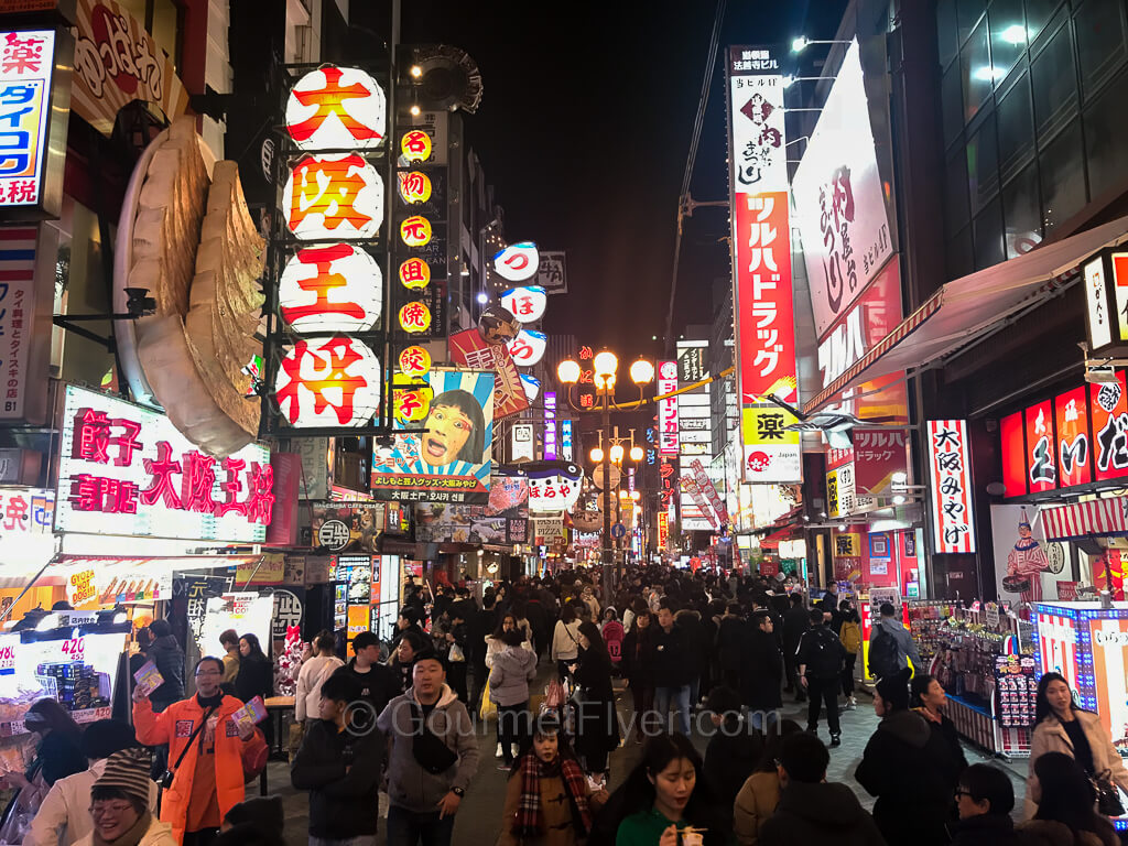 Moderately priced iconic cultural places to eat in Osaka is shown in the heart of Dotonbori where huge crowds roam the streets under bright neon signs.