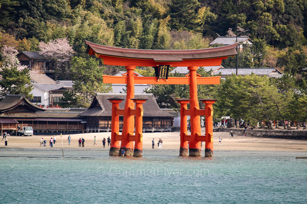 A large torii gate stands on the water with a beach in the background.