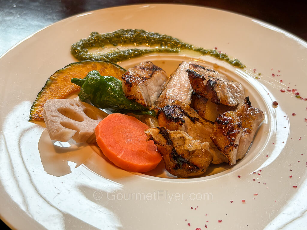 A dish of roasted pork is served with a blend of vegetables and garnished with sauces on the plate.