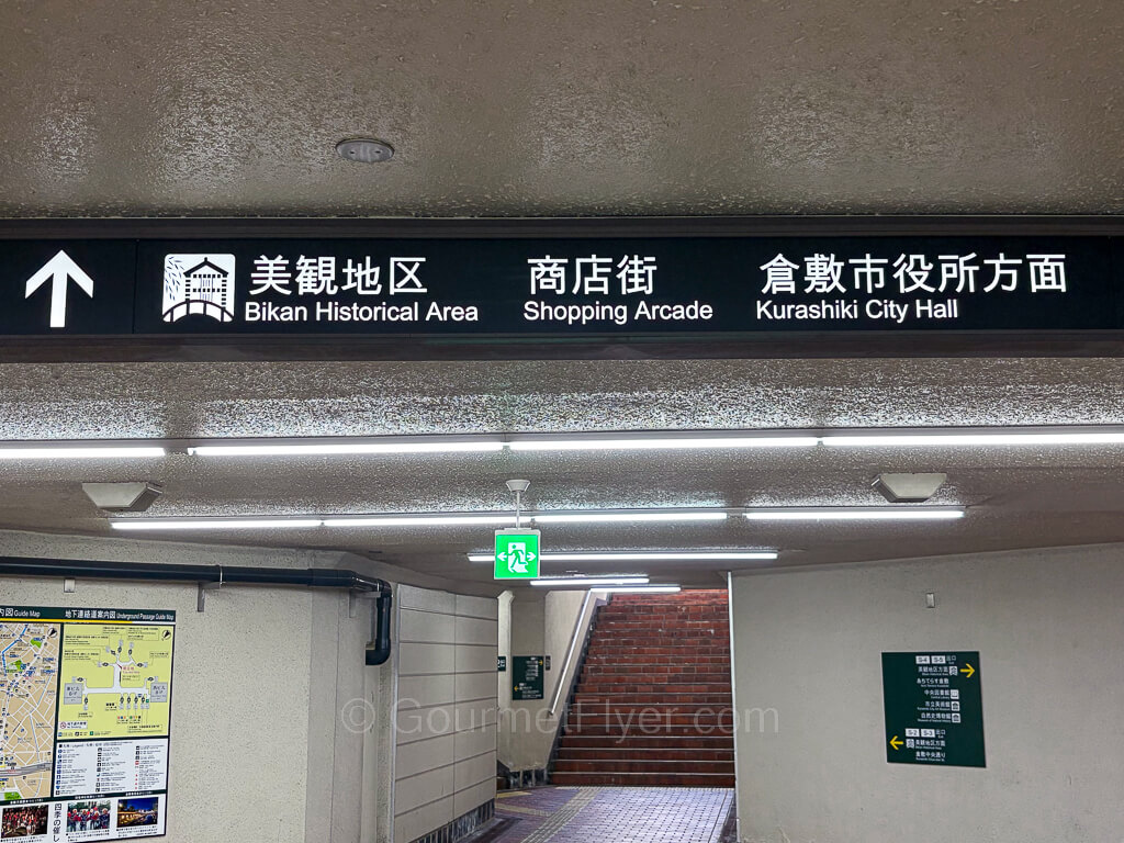 Lighted signs with white words in black background hang from the roof of an underground tunnel.