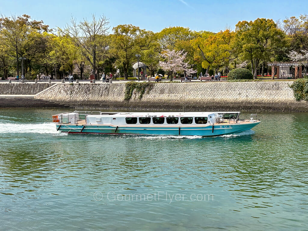 A small ferry sails along a canal lined with trees with thick green leaves.